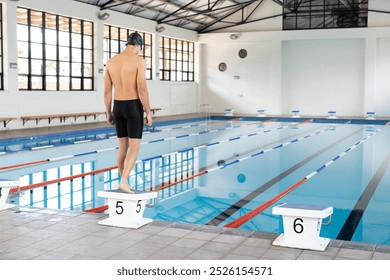 Caucasian young male swimmer standing on starting block at pool indoors, copy space. He has short dark hair, wearing black swimwear, preparing for training, unaltered - Powered by Shutterstock