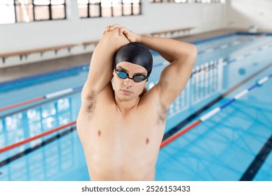 Caucasian young male swimmer stretching before swimming indoors, pool in background. He has short brown hair, wearing goggles, and looks focused, unaltered. - Powered by Shutterstock