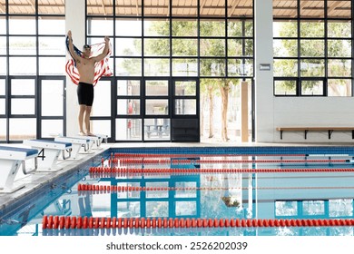 Caucasian young male swimmer stretching on pool edge indoors, holding american flag, copy space. He has short brown hair, wearing swim cap and goggles, and striped towel, unaltered - Powered by Shutterstock