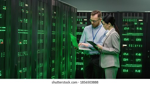 Caucasian young man analytic with laptop computer in servers room talking with woman colleague. Male and female co-workers of database center talking and discussing security. Monitoring concept. - Powered by Shutterstock