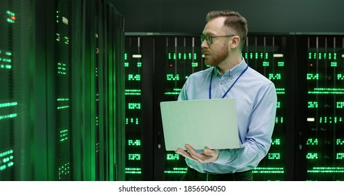 Caucasian young man in glasses, software specialist walking at servers with laptop computer, typing on keyboard and checking big data processors. Male analytic working on digital information security.