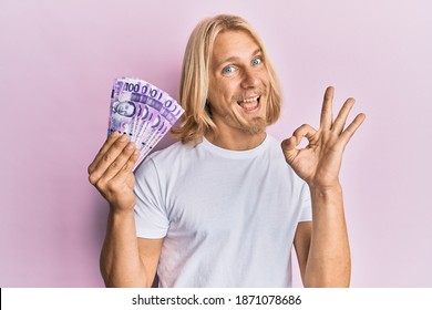 Caucasian young man with long hair holding 100 philippine peso banknotes doing ok sign with fingers, smiling friendly gesturing excellent symbol 