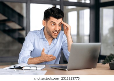 Caucasian young man, manager, freelancer or office worker, sitting at table in office, using laptop, shocked by news or message, looking at screen in surprise, gesturing with his hand, puzzled - Shutterstock ID 2311823775