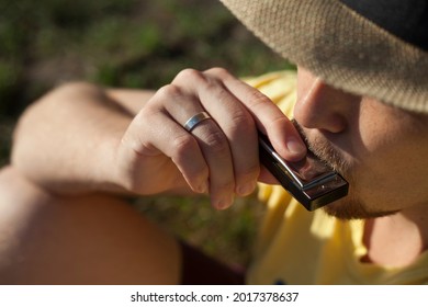 Caucasian young man in straw hat playing harmonica outdoors. Rural slow life. Farmer with mustache playing music in the field. Man holds musical instrument in hands closeup with selective focus.