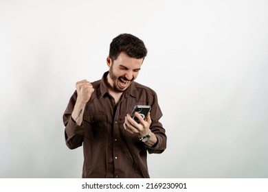 Caucasian Young Man Wearing Shirt Posing Isolated Over White Background Being Happy Winning A Bet In Online Sport Gambling Application On His Mobile Phone.