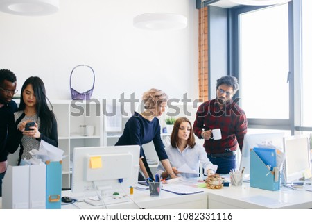 Caucasian young manager showing the documents to beautiful woman with fair hair and Hindu man holding tea in the office with panorama window