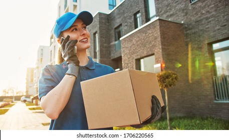 Caucasian young pretty woman, delivery worker in blue hat cap walking the street, carrying cardbox parcel and talking on mobile phone. Female beautiful courier with carton box speaking on cellphone.