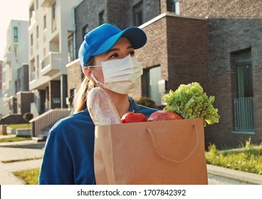 Caucasian Young Pretty Woman, Delivery Worker In Bluetooth Headphones, Cap And Medical Mask Walking The Street And Carrying Packet With Fresh Food. Female Beautiful Courier Going To Deliver Grocery.