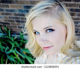 Caucasian Young Woman With Blonde Hair And Blue Eyes Looks At Camera For Selfie In Front of Brick Wall and Greenery - Powered by Shutterstock