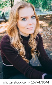 Caucasian, young woman with blue eyes and long, blonde hair wearing maroon crop top and black jeans. Smiling on park bench.