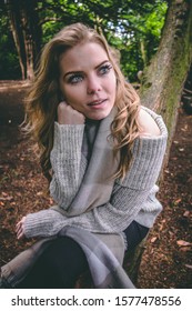 Caucasian, Young Woman With Blue Eyes And Long, Blonde Hair Wearing Off-the-shoulder Grey Sweater And Fuzzy, Checkered Scarf And Black Jeans. Daydreaming While Leaning On Her Face On Tree.