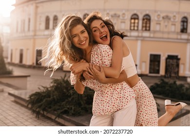 Caucasian young woman brunette hung on back of her friend against the open background of city. Girls are smiling broadly with their teeth at camera, dressed in white outfits.