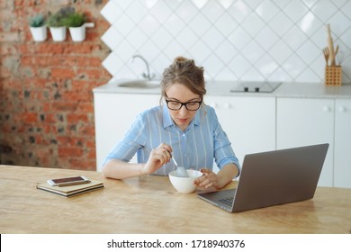 Caucasian young woman with dark blond hair in glasses and blue striped shirt sits in bright kitchen with brick wall and working or studying on her laptop during breakfast. Watching tv series or movie