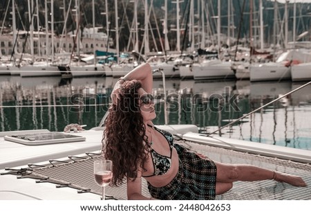 caucasian young woman, dark curly long hair drinking rose wine on front deck of white sailing catamaran in marina at berth