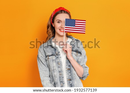 Caucasian young woman in denim jacket covers half of her face with a small american flag and smiles isolated over orange background, student holding USA flag, 4th of july independence day, copy space