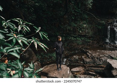caucasian young woman in disheveled black jacket standing on big rock with hands in pockets smiling calm and relaxed admiring beauty of waterfalls,purakaunui falls, new zealand - Shutterstock ID 2245070747