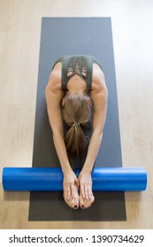 Caucasian young woman doing typical stretching, head to knees with foam blue roller, paschimottanasana pose, indoor at club, front view, selective focus. Yoga, pilates, stretching, exercise therapy