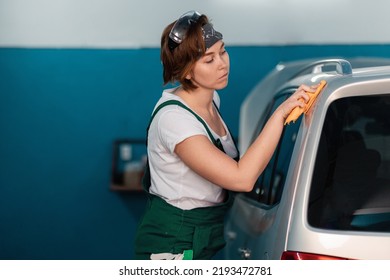 Caucasian Young Woman In A Green Coveralls Wiping A Car With A Rag. Work In An Auto Repair Shop And Garage.