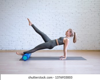 Caucasian Young Woman In Grey Sports Suit Doing Pilates Workout On Math With Roller, Loft Style Background, Toned Light Blue. Wellness, Recovery, Rehabilitation, Prevention Spine Desease, Smart Body