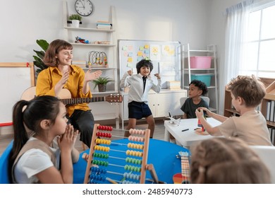 Caucasian young woman teacher playing guitar with students in school. Adorable little children having fun enjoy education activity, dance with instrument in classroom at preschool nursery kindergarten - Powered by Shutterstock