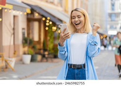 Caucasian young woman using mobile smartphone celebrating win good message news, lottery jackpot victory, giveaway online outdoors. Happy adult lady tourist standing in city street. Town lifestyles - Powered by Shutterstock