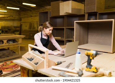 Caucasian Young Woman Working On A Laptop Inside A Woodshop. Female Worker Sitting In A Table Surrounded By Carpentry Tools And Furniture