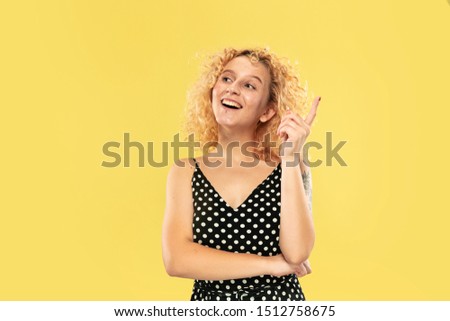 Caucasian young woman's half-length portrait on yellow studio background. Beautiful female model in black dress. Concept of human emotions, facial expression. Smiling, having an idea, laughting.