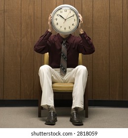 Caucasion Mid-adult Retro Businessman Sitting In Chair Against Wood Paneling Holding Clock Over Face.