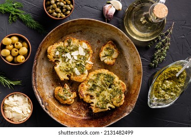 Cauliflower steak with spices lies in a frying pan. Olive oil, chimichurri sauce, capers, olives, herbs, various spices side by side. Dark background. Vegetarian food.