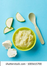 Cauliflower And Zucchini Baby Puree In Bowl With Baby Spoon On Blue Background, Top View