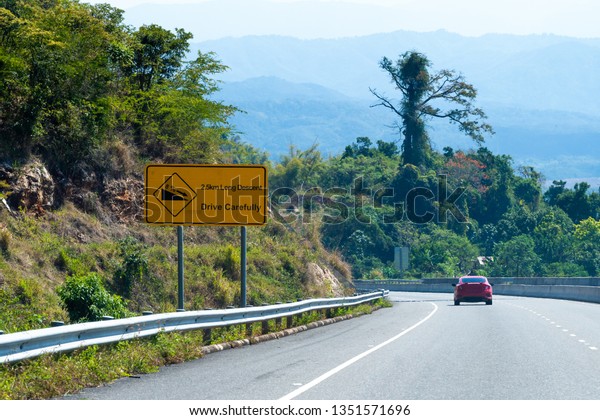 Caution Long Descent 2.5km downhill road sign on\
highway through mountains in scenic countryside. Red sedan/coupe\
sports car driving on left hand side on 8% decline down the hill on\
two lane street.