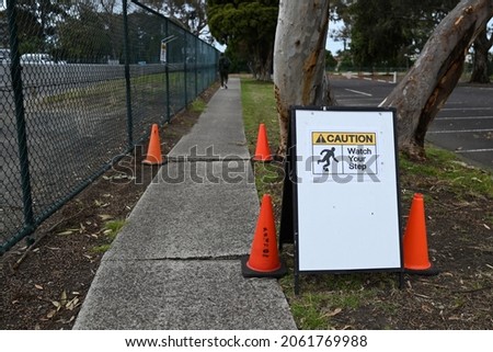 A caution watch your step sign, warning about uneven pavement on a footpath, with orange traffic cones. An out of focus person can be seen walking the path in the background