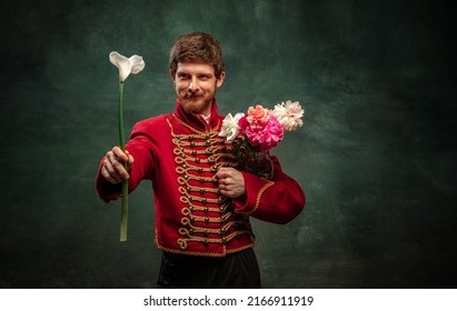 Cavalier and beau. Handsome young man in image of medieval hussar holding flowers isolated on dark blue background. Retro style, comparison of eras concept. Male model wearing hussar uniform. - Shutterstock ID 2166911919