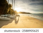 Cavaliere Beach at sunrise with golden light, star-shaped sun and blue sky. Catamarans on sand with pine trees behind. Foam in the waves and distant hills. In le Lavandou, Var department, Provence.