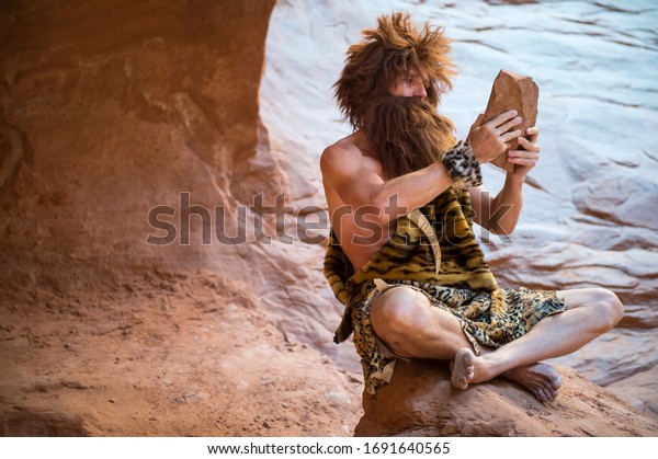 Caveman watching the screen of his\
primitive stone tablet outdoors in a weathered rock\
cave