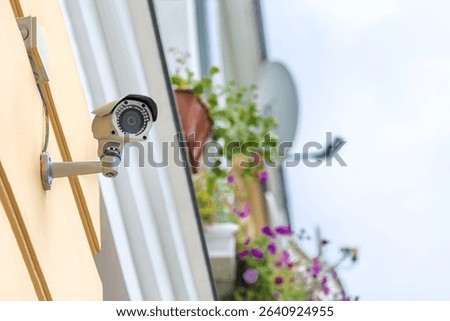 The CCTV camera is installed on the facade of a residential building. In the background is a balcony with flowers and a satellite dish. Ideal for security and video surveillance topics.