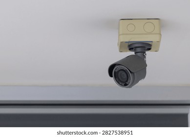 CCTV security camera mounted on ceiling. Gray dome lens faces downward. Beige housing with two circular buttons. Clean, neutral wall background. Ideal for security, surveillance, or safety themes