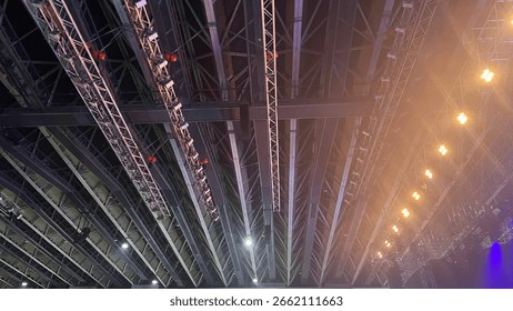 Ceiling view of a large indoor arena featuring a complex metal truss system with professional stage lighting