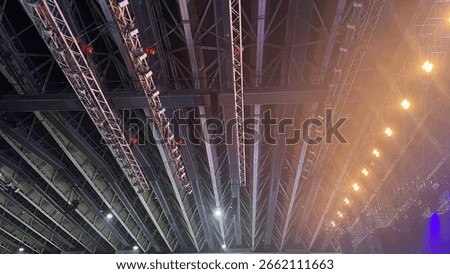 Ceiling view of a large indoor arena featuring a complex metal truss system with professional stage lighting