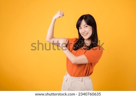 Celebrate with confidence as a young Asian woman 30s showcases a fist up hand sign gesture, wearing an orange shirt on yellow background. Empowerment and feminism concept.