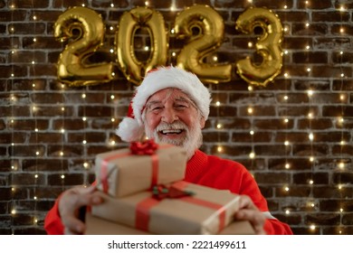 Celebrate Event New Year For Caucasian Bearded Elderly Male Holding Christmas Presents - Beautiful Senior Adult Man In Santa Hat Celebrate Holiday At Home By Night.