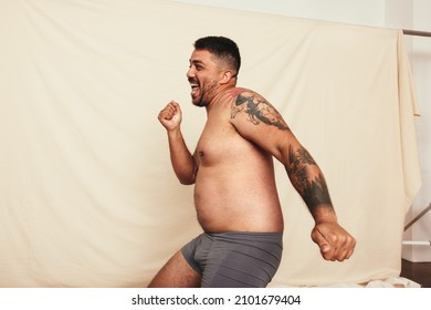 Celebrating being natural. Happy shirtless man dancing cheerfully while wearing grey underwear and standing against a studio background. Body positive and self-confident man embracing his own body.