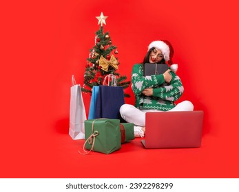 Celebrating Christmas alone concept, woman full body sit floor red background near Christmas tree hugging present box using laptop smiling feel bittersweet happiness. Shopping bags, gift boxes. - Powered by Shutterstock