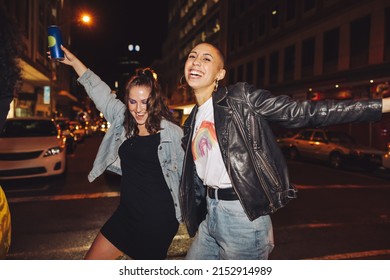 Celebrating With Dance. Two Happy Female Friends Dancing Cheerfully While Crossing A Street In The City. Two Vibrant Young Women Laughing And Having A Good Time On A Weekend Night.