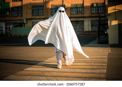 Celebrating halloween. Ghost Challenge 2021. A man dressed as a ghost from a sheet and sunglasses crosses the road along the crosswalk. Spooky season. 