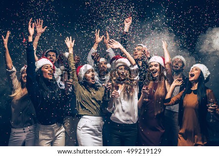 Celebrating New Year together. Group of beautiful young people in Santa hats throwing colorful confetti and looking happy