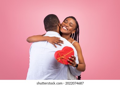 Celebrating Special Occasion. Portrait Of Romantic African American Guy Giving His Girlfriend Wrapped Box With Present In Heart Shape And Hugging, Man Greeting Lady With Holiday, Pink Studio Wall