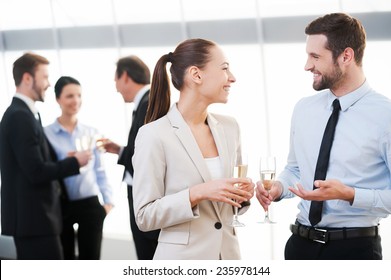 Celebrating Their Common Success. Two Cheerful Business People Drinking Champagne And Talking While Other People Communicating In The Background 