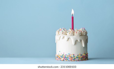Celebration birthday cake decorated with white drip icing, buttercream frosting swirls, colorful sugar sprinkles and one birthday candle against a plain blue background