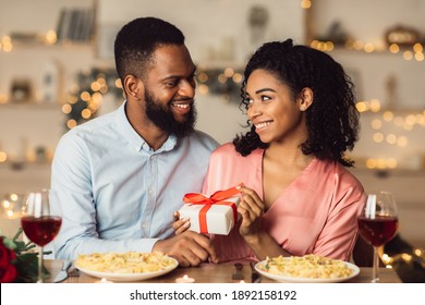 Celebration Concept. Happy Black Young Woman Unwrapping Box With Gift, Pulling Red Ribbon And Looking At Her Man With Love And Appreciation, Receiving Present. African Couple Having Romantic Date